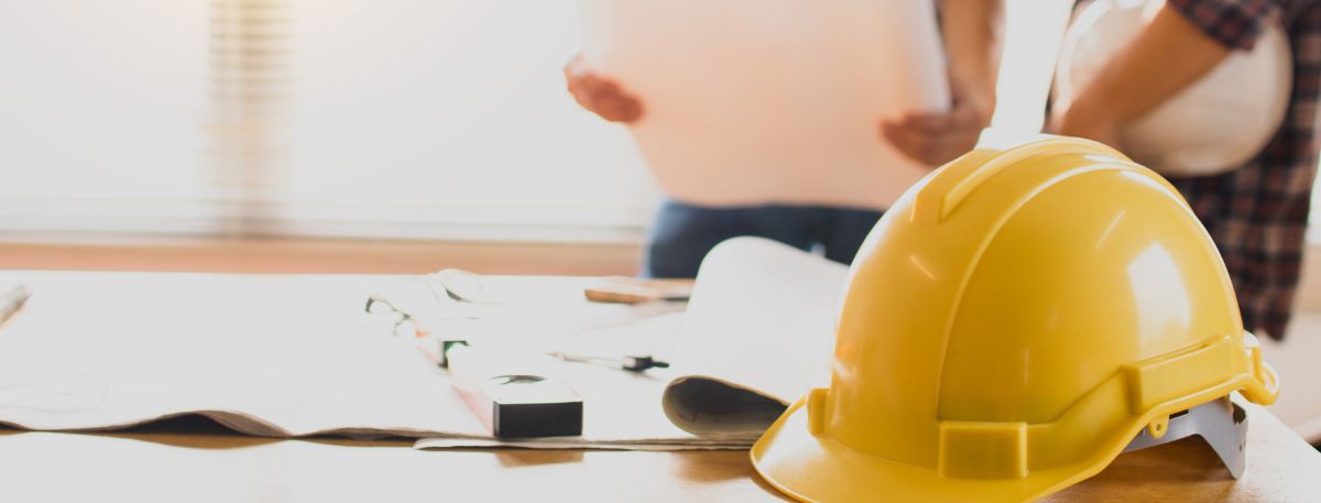 A yellow hard hat on a table with some building plans and people in the background