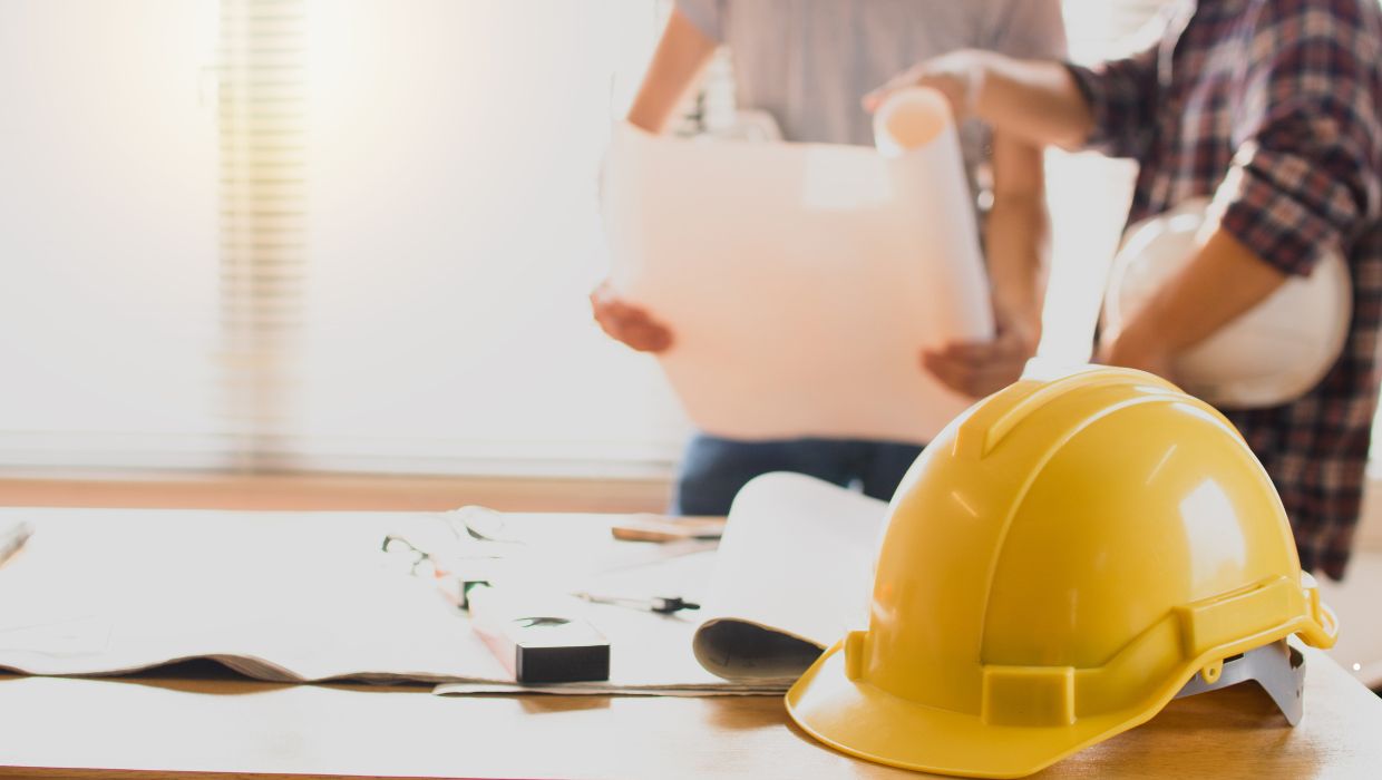 A yellow hard hat on a table with some building plans and people in the background tile